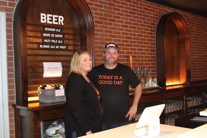 Two people standing at a bar with a brick wall and beer sign in the background
