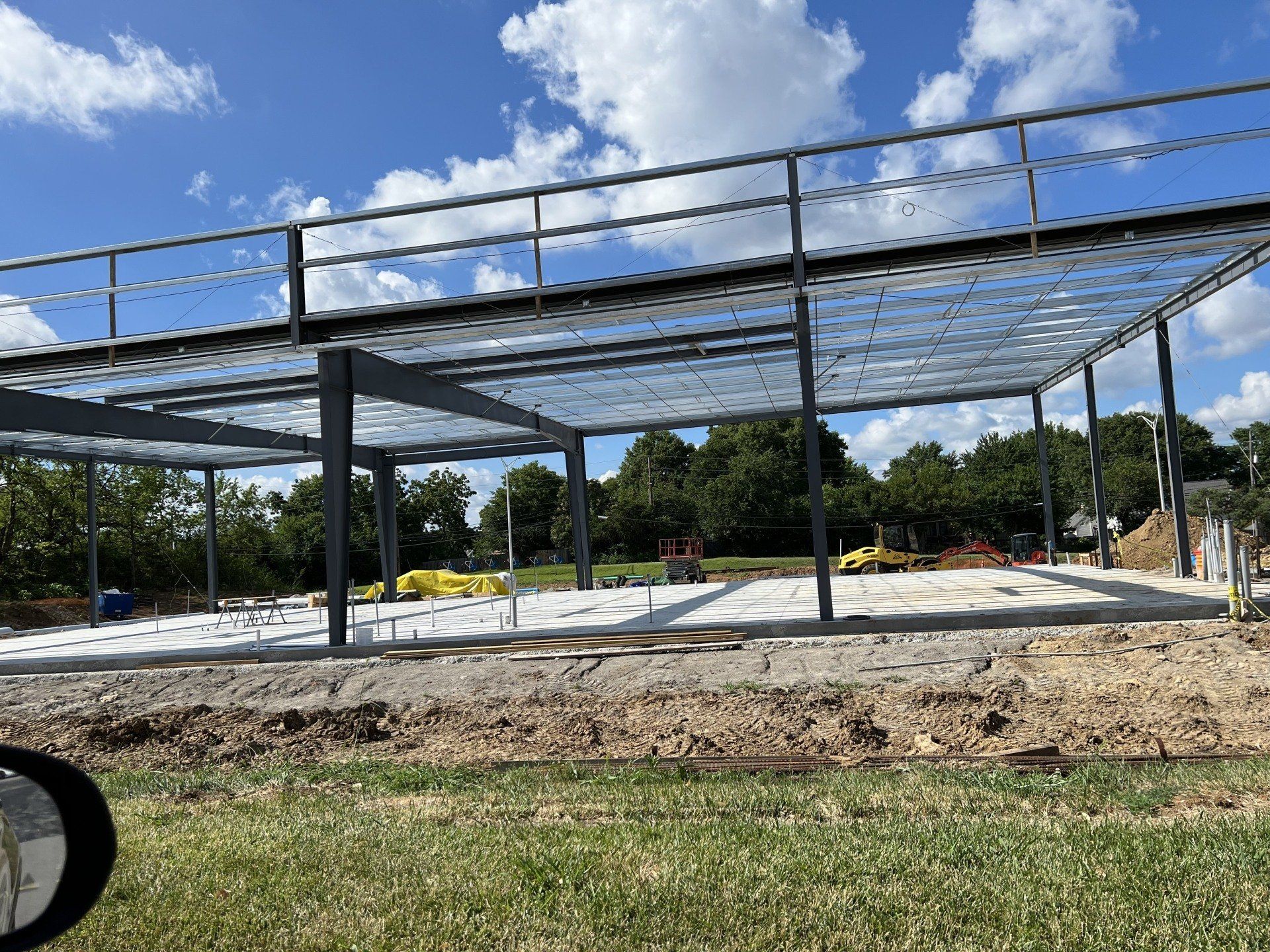 Steel-frame building under construction on a dirt lot, with concrete pads and grass in the foreground.