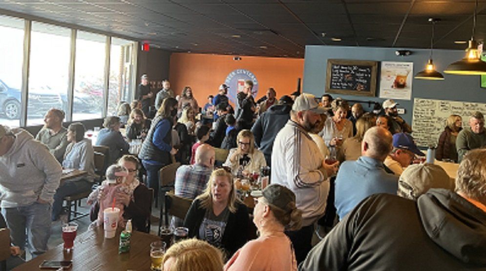 Crowded restaurant with people seated and standing, chatting near large windows and a warm orange wall.