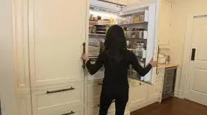 Woman in black outfit opening a large, built-in refrigerator in a light-colored kitchen.