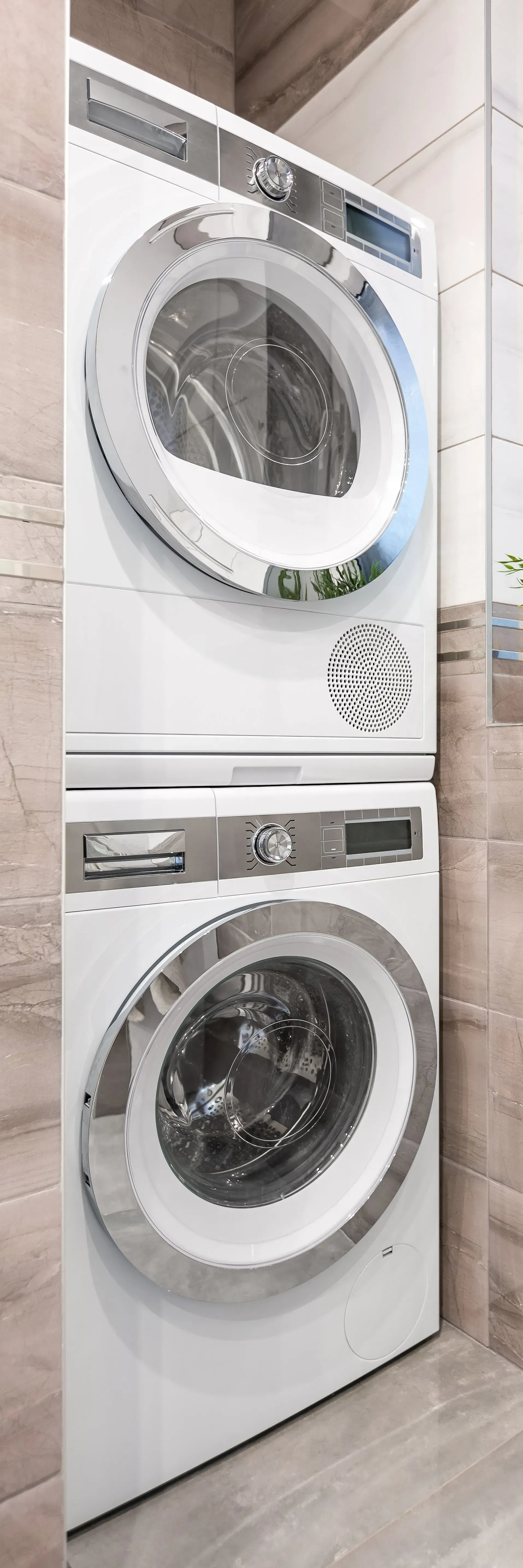 A stacked washer and dryer unit in a small bathroom, white with chrome accents, positioned against a tiled wall and floor.