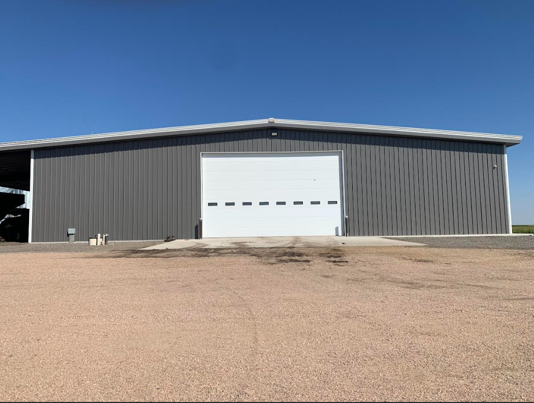Large gray metal building with a white garage door under a blue sky, viewed across a gravel lot