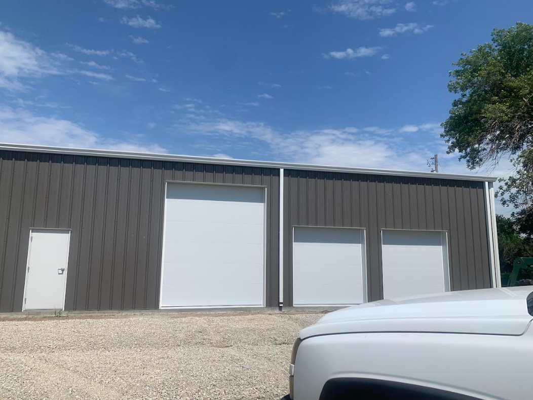 Gray industrial warehouse with large garage doors under a blue sky, with a white vehicle in the foreground
