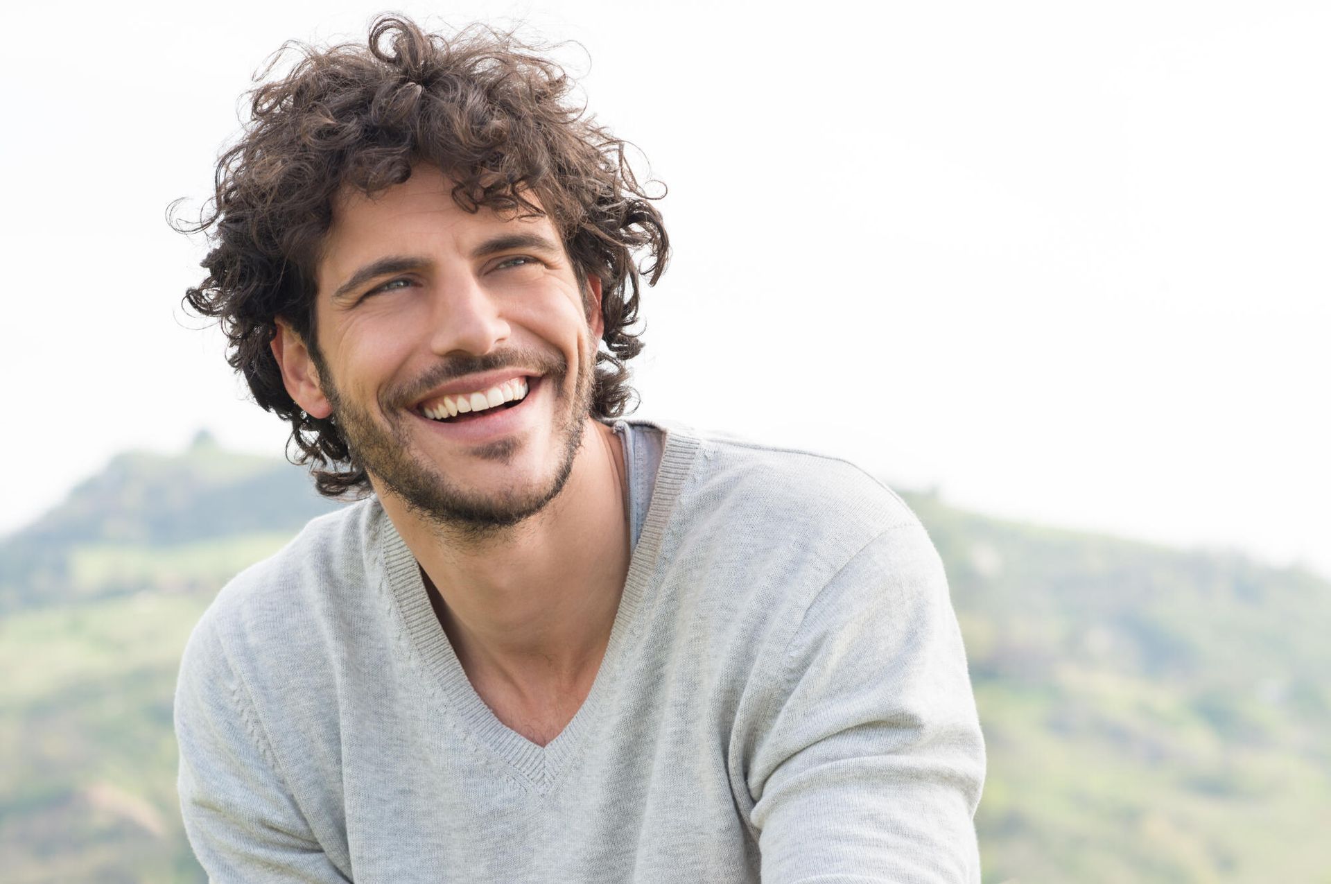 Man with curly brown hair smiles, wearing a gray sweater, outdoors with a blurry green background.
