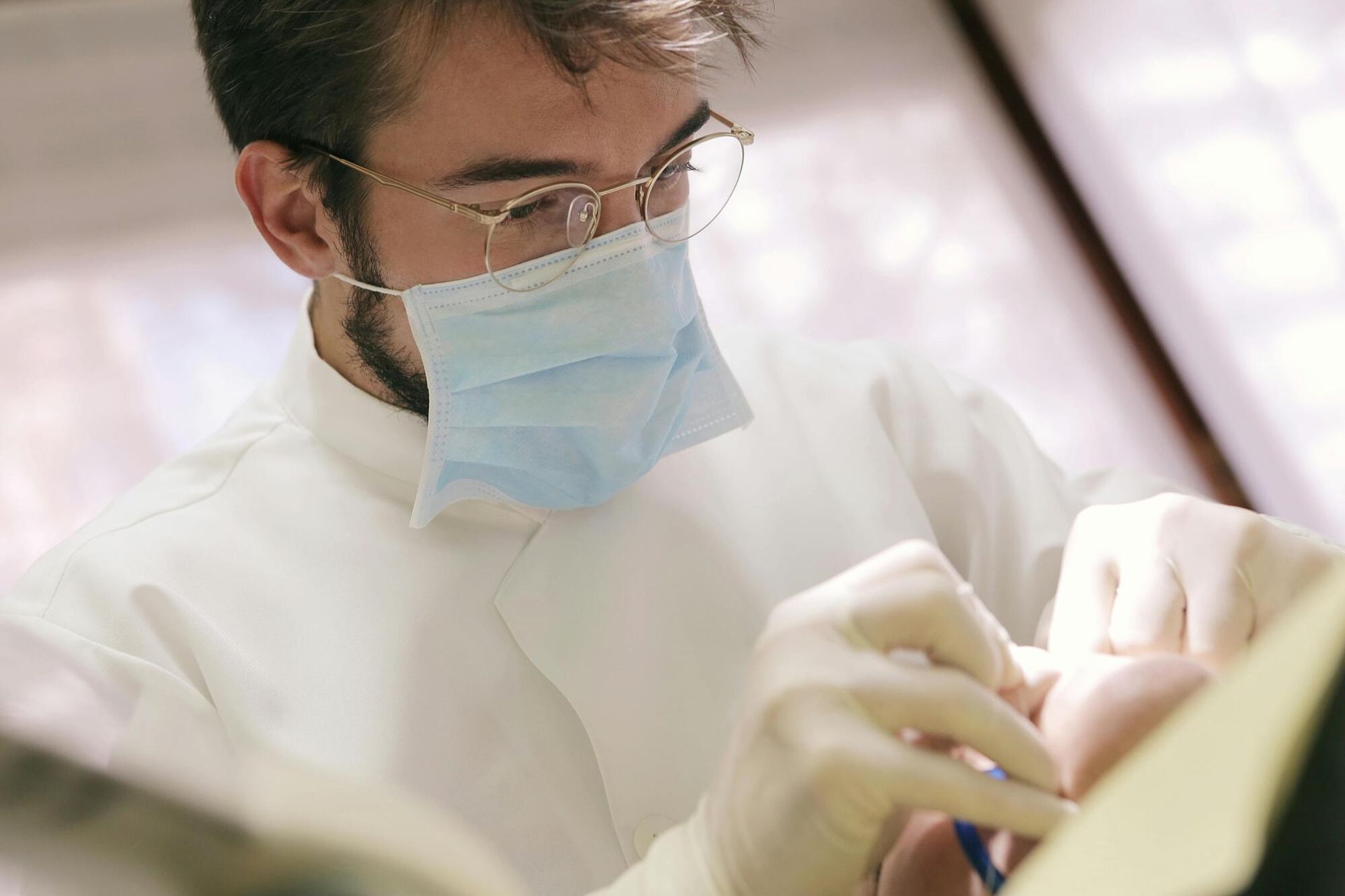 Dentist examining a patient's mouth. He wears glasses, a mask, and gloves, in a dental office.