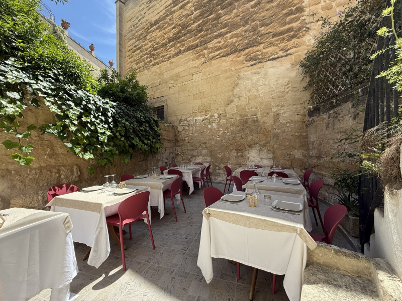 Terrazza esterna del ristorante con tavoli bianchi e sedie rosa accanto a muri in pietra e vegetazione.
