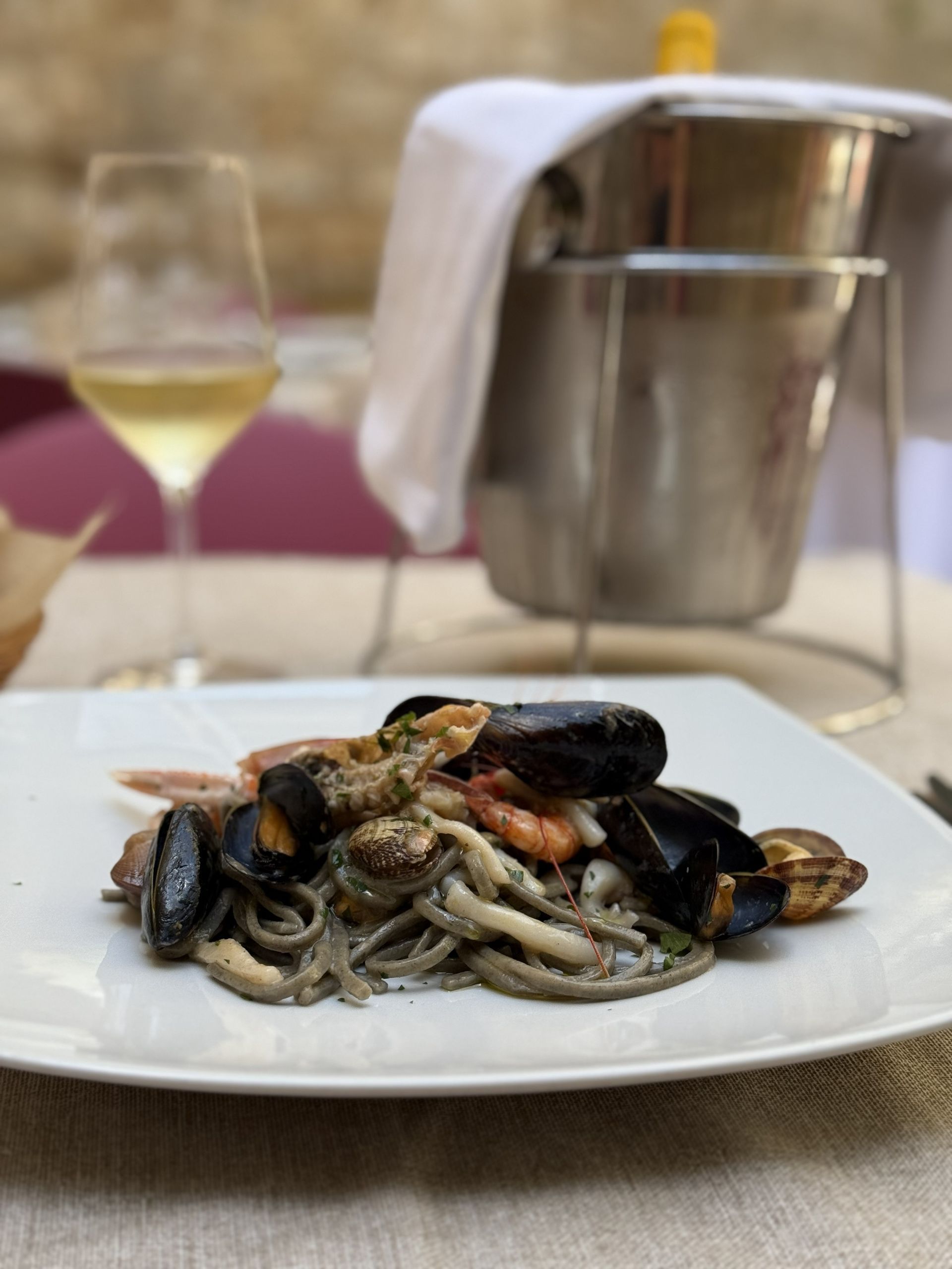 Un piatto di pasta con cozze e calamari su un tavolo, con un bicchiere di vino e un secchiello del ghiaccio sullo sfondo.