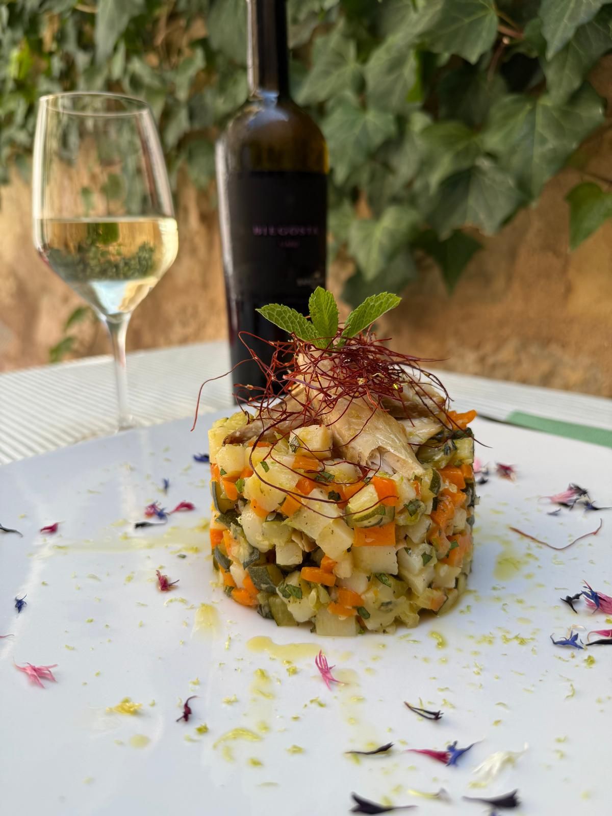 Insalata gourmet di verdure a cubetti con menta su un piatto bianco, accanto a un bicchiere di vino bianco e una bottiglia di vino all'aperto.