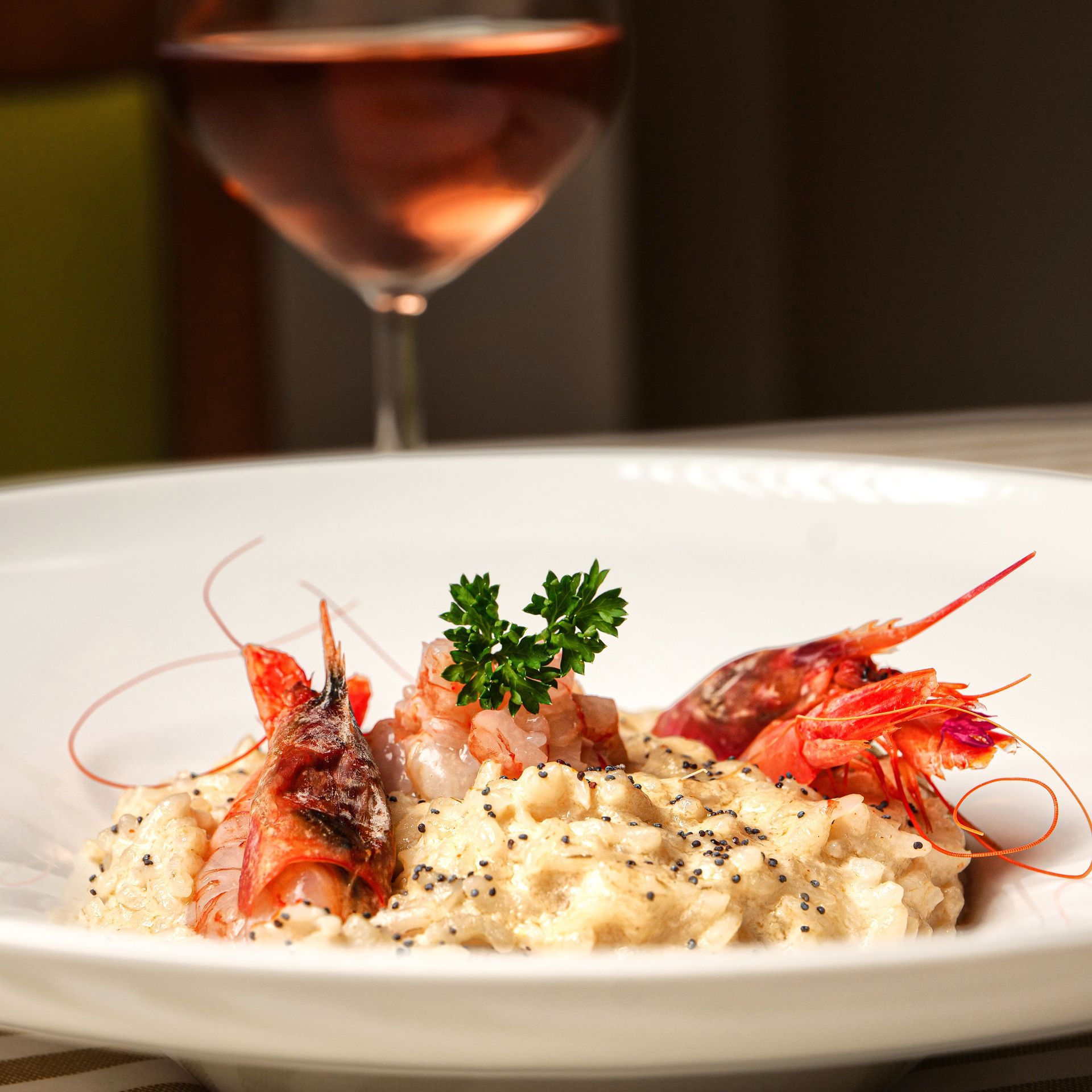 Risotto ai gamberi guarnito con prezzemolo su un piatto bianco, con un bicchiere di rosé sullo sfondo.