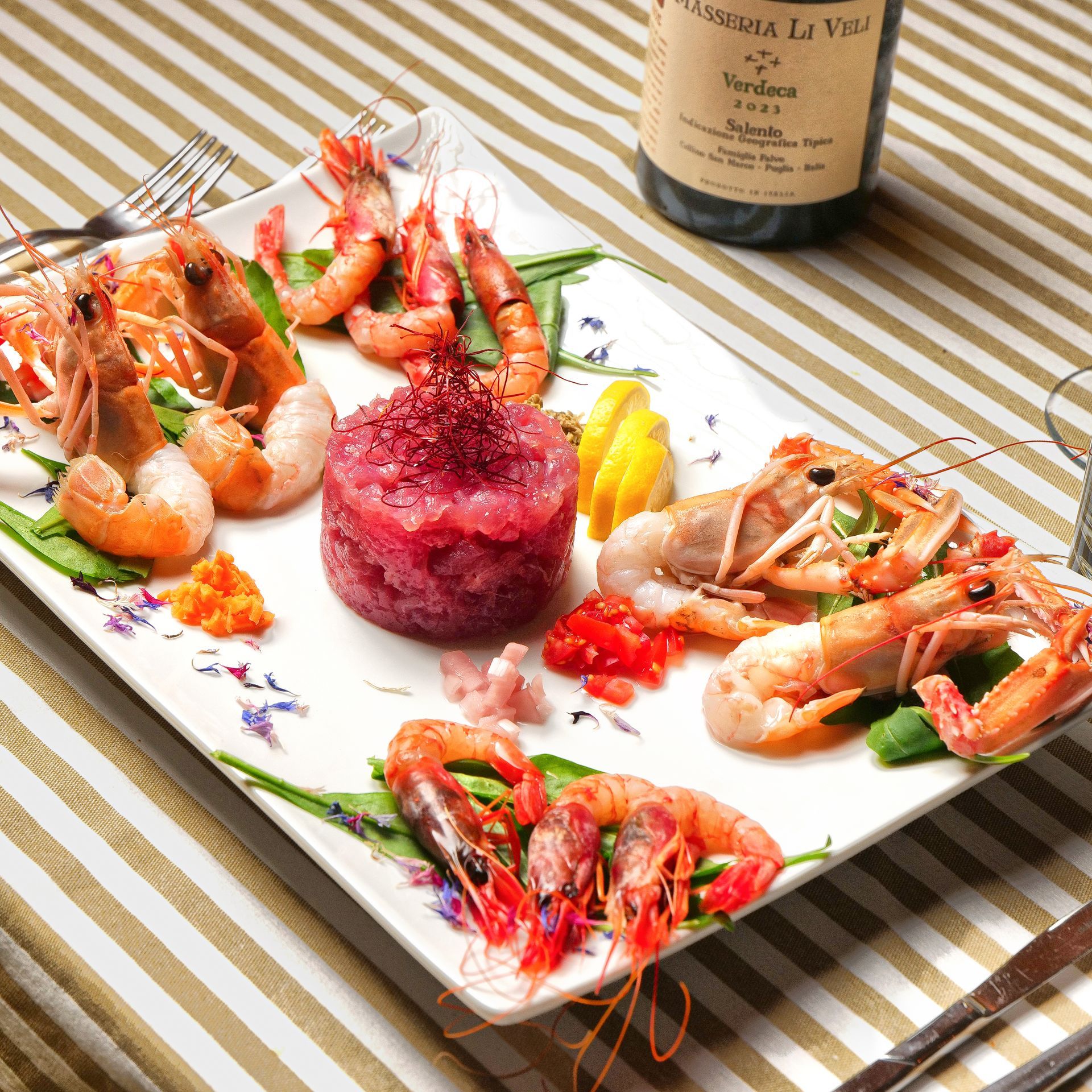 Piatto di frutti di mare con gamberi, scampi e un mucchietto di riso rosso su un vassoio bianco decorato con fiori, accanto a una bottiglia di vino.