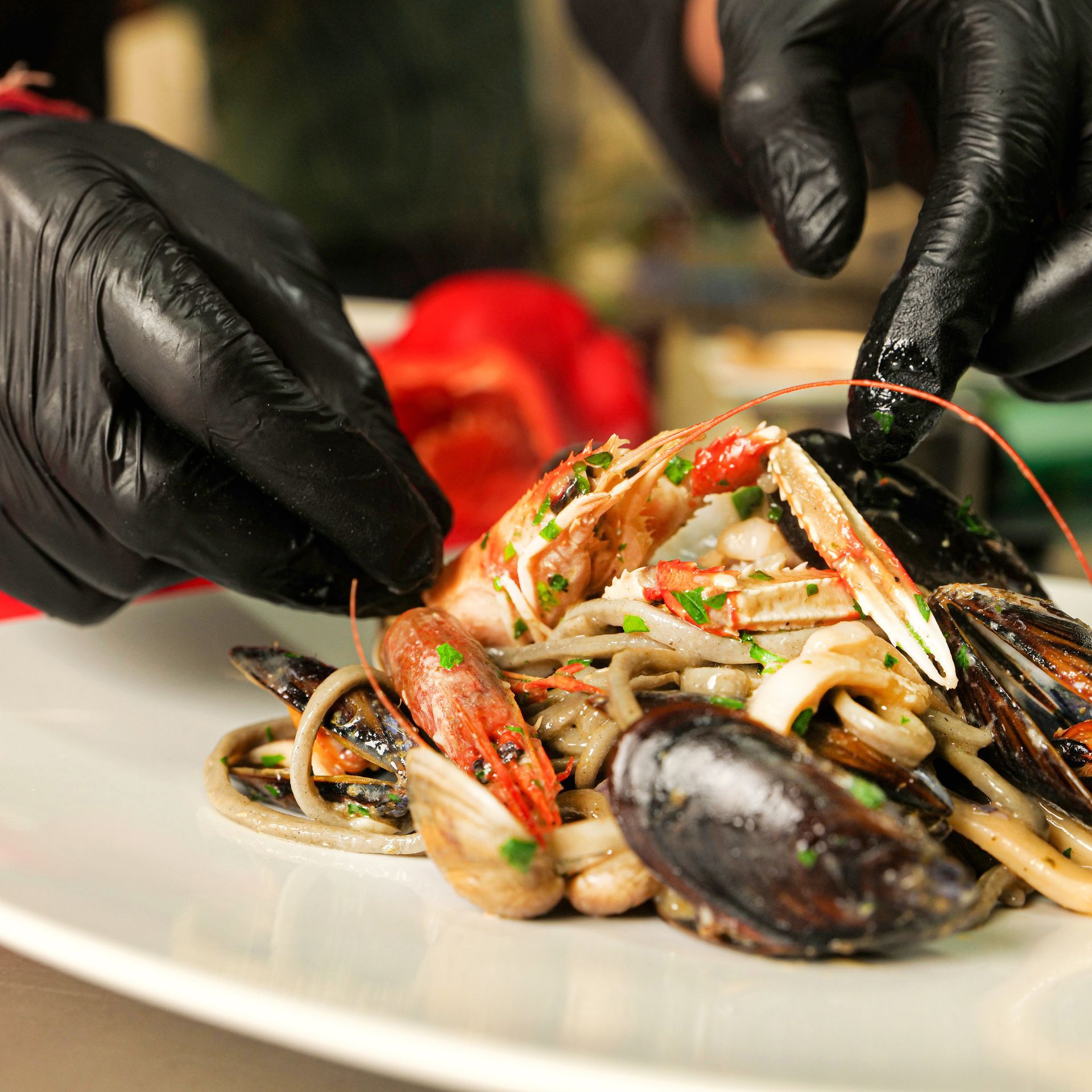 Mani guantate di nero impiattano pasta ai frutti di mare con cozze e gamberi su un piatto bianco.
