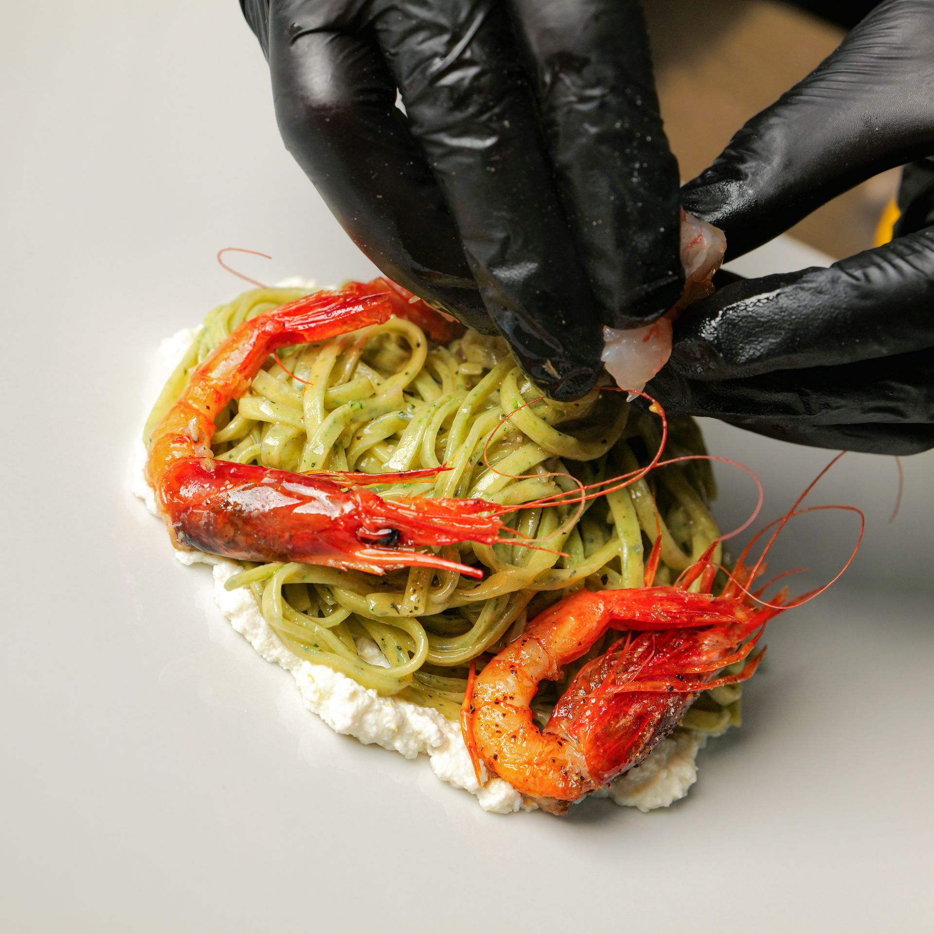 Mani guantate che impiattano pasta verde con gamberi rossi su un piatto bianco.