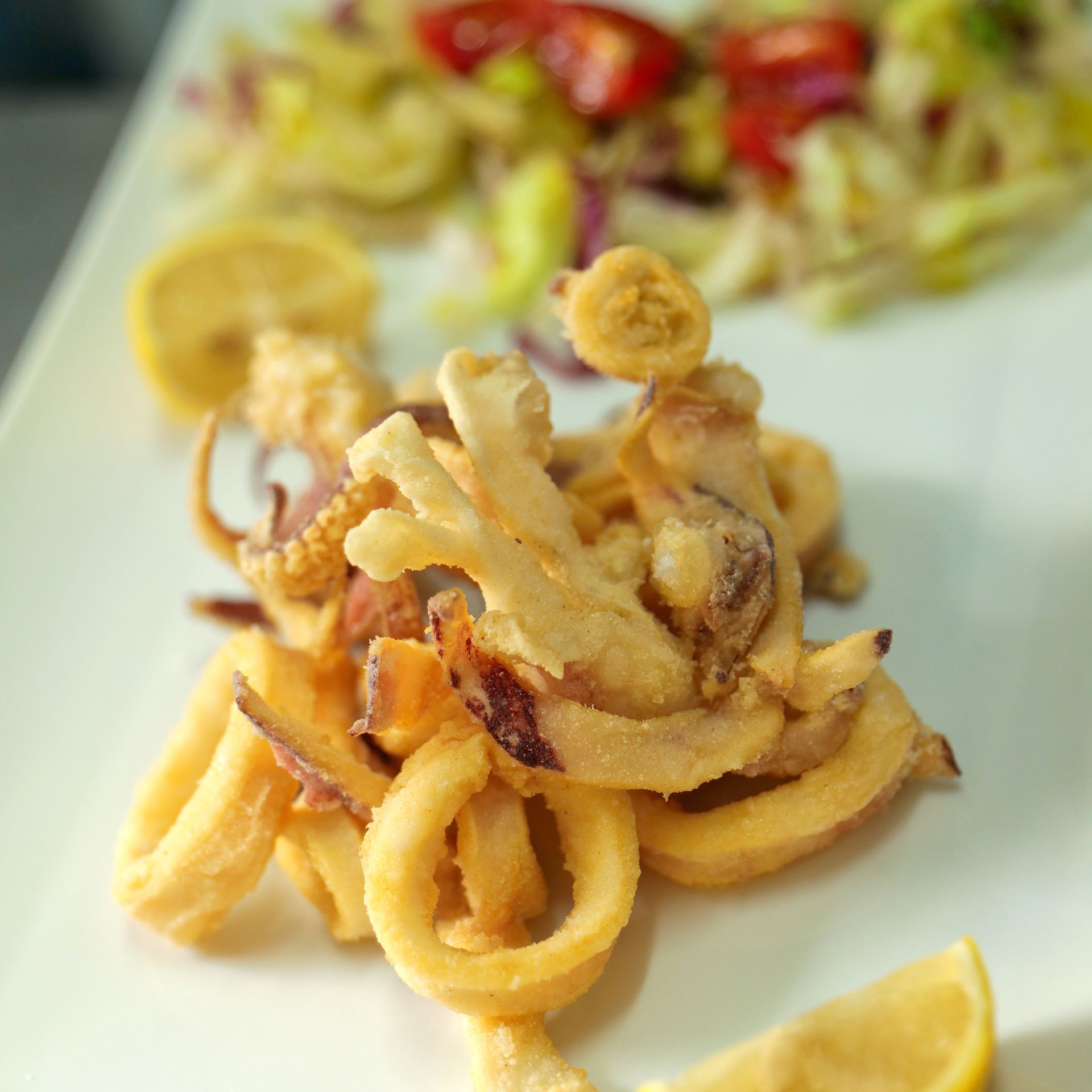 Anelli di calamari fritti con spicchi di limone su un piatto, con insalata sullo sfondo.