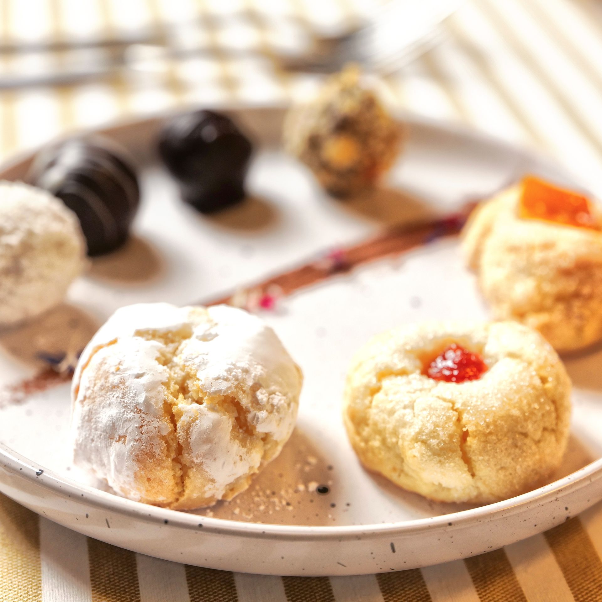 Su un piatto bianco, un assortimento di biscotti e pasticcini ricoperti di zucchero a velo, con un biscotto ripieno di marmellata in primo piano.