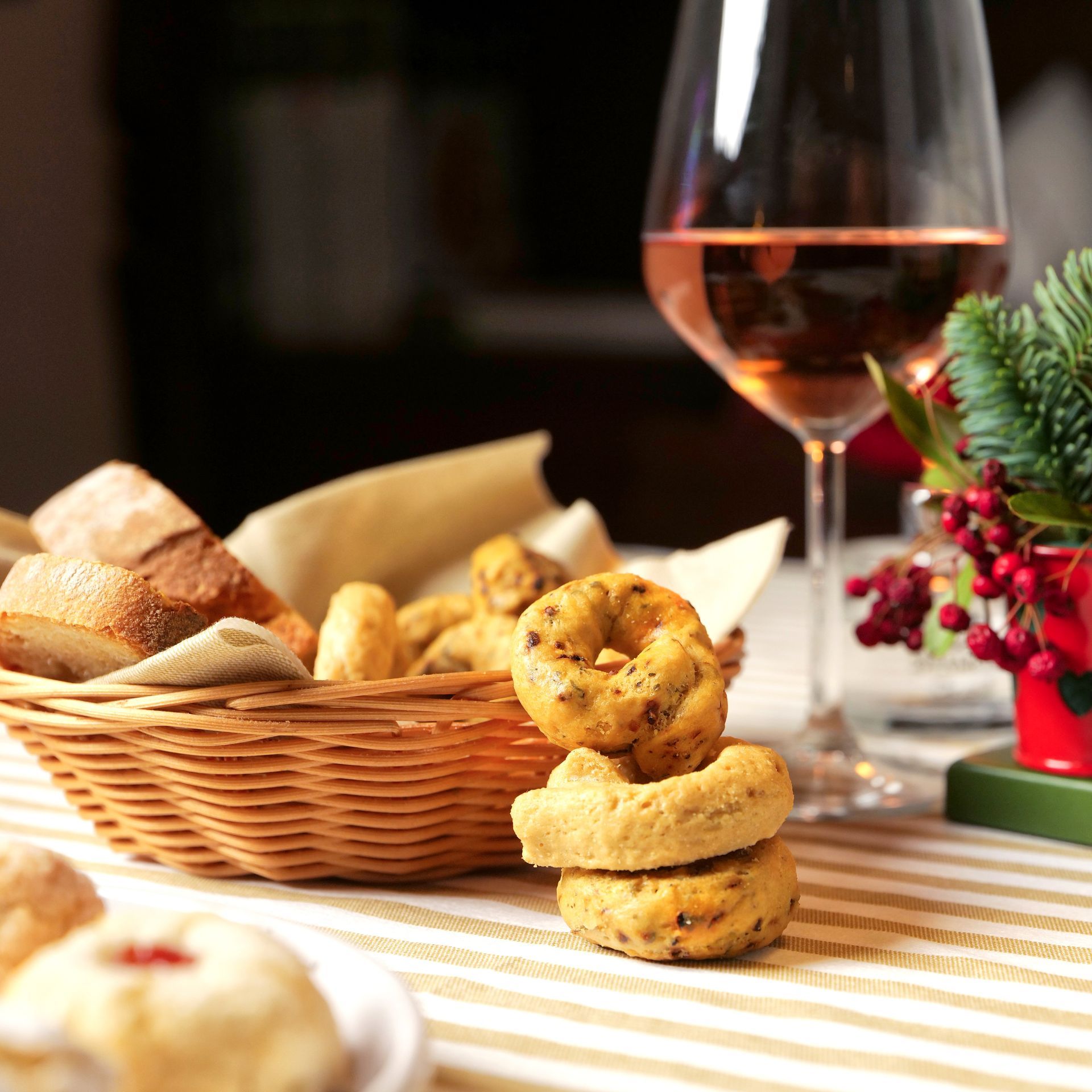 Un cestino di pane e biscotti accanto a un bicchiere di rosé su una tovaglia a righe, con verde e frutti di bosco.