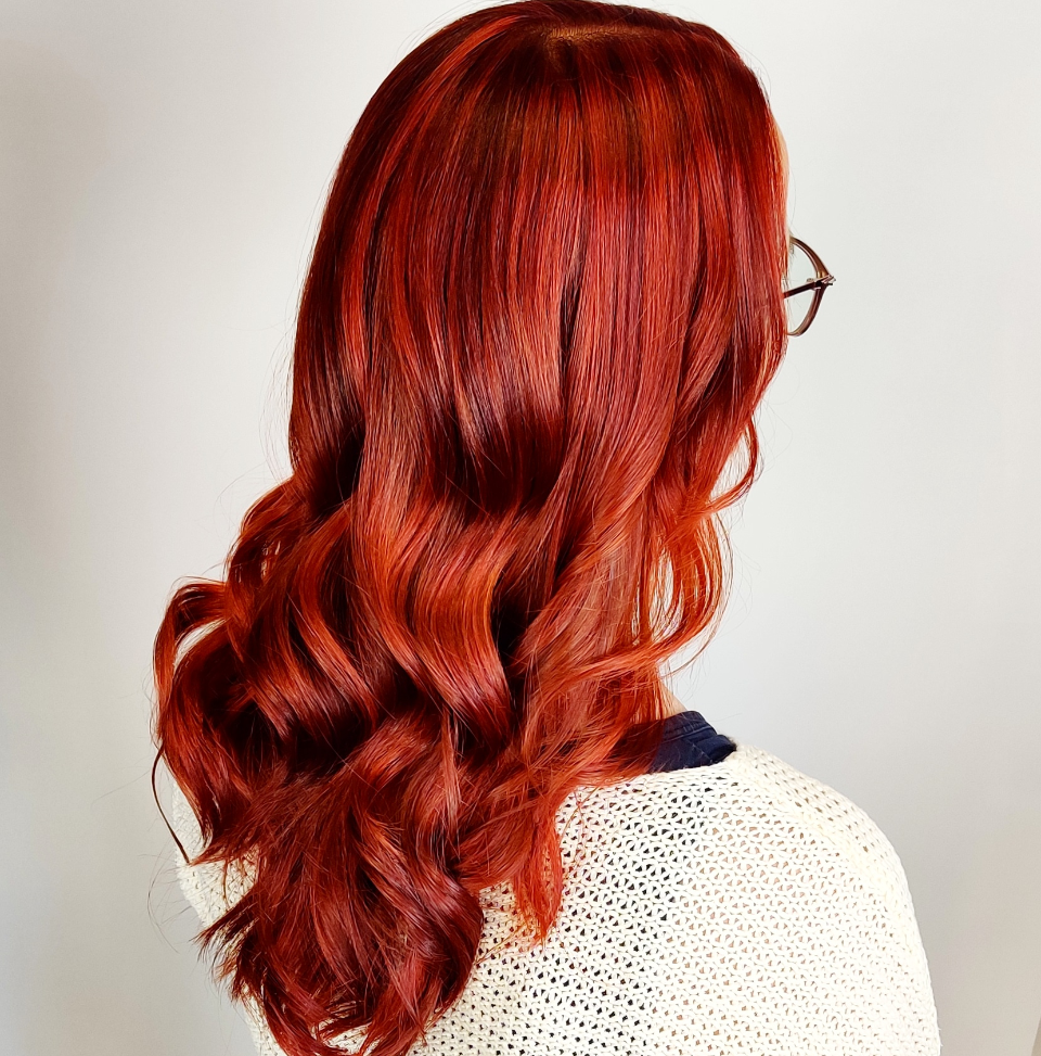 Woman with long, wavy red hair wearing glasses and a white sweater against a light gray wall.
