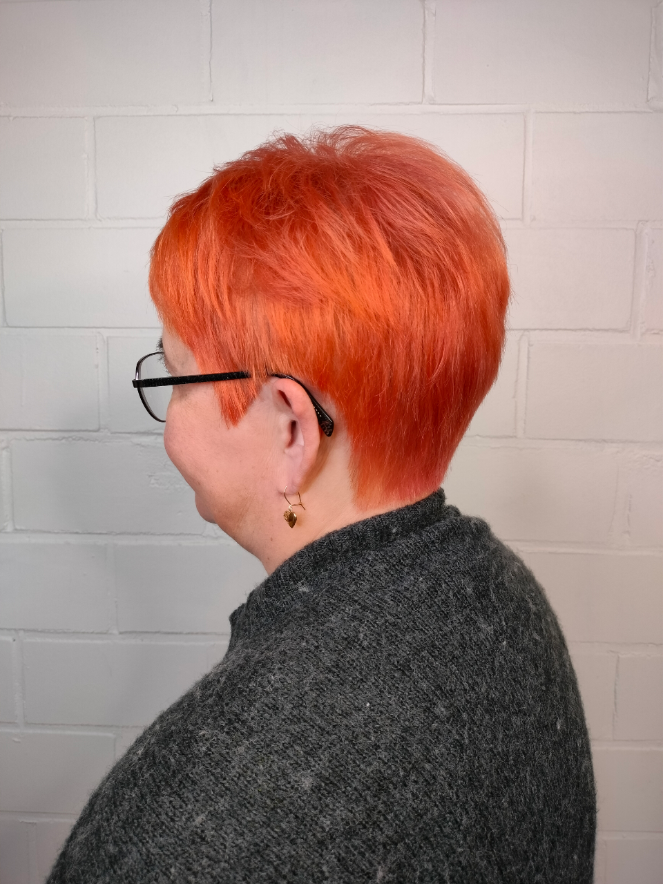 A woman with short red hair and glasses is wearing a gray sweater.