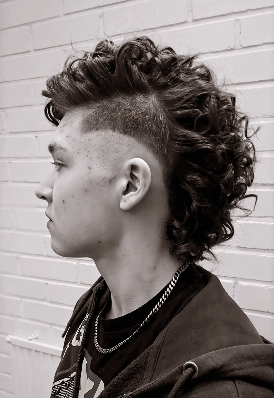 A young man with curly hair and a mohawk hairstyle is standing in front of a brick wall.