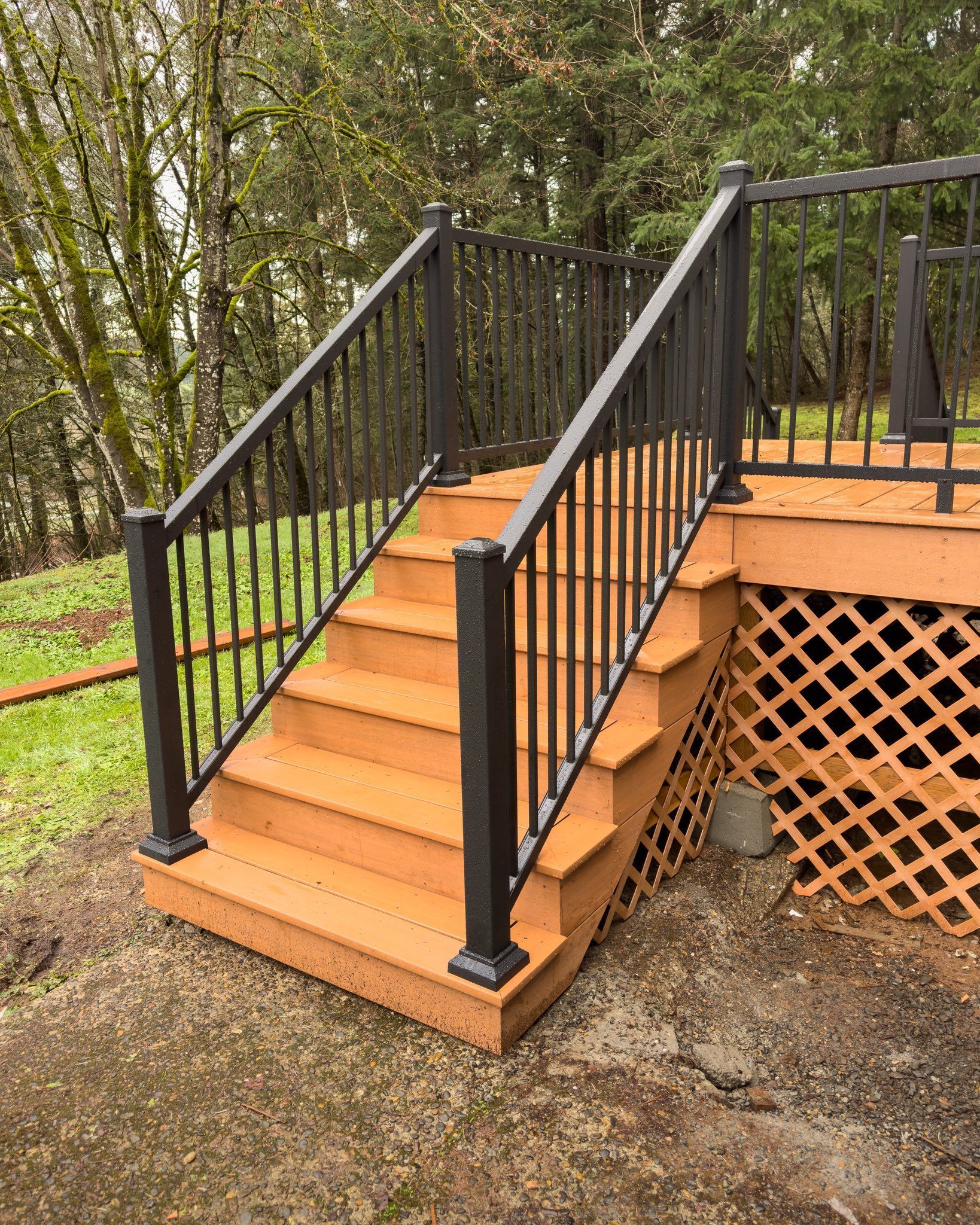 Wooden deck stairs with black metal railings leading up to a deck with lattice skirting, set against a wooded background.