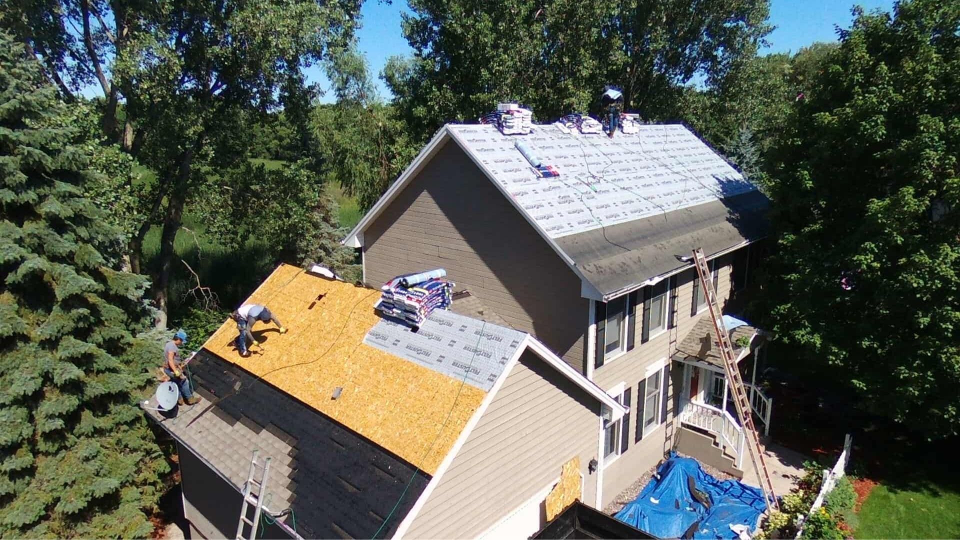 Construction workers repairing a two-story residential roof, with sections showing bare wood, underlayment, and shingles.