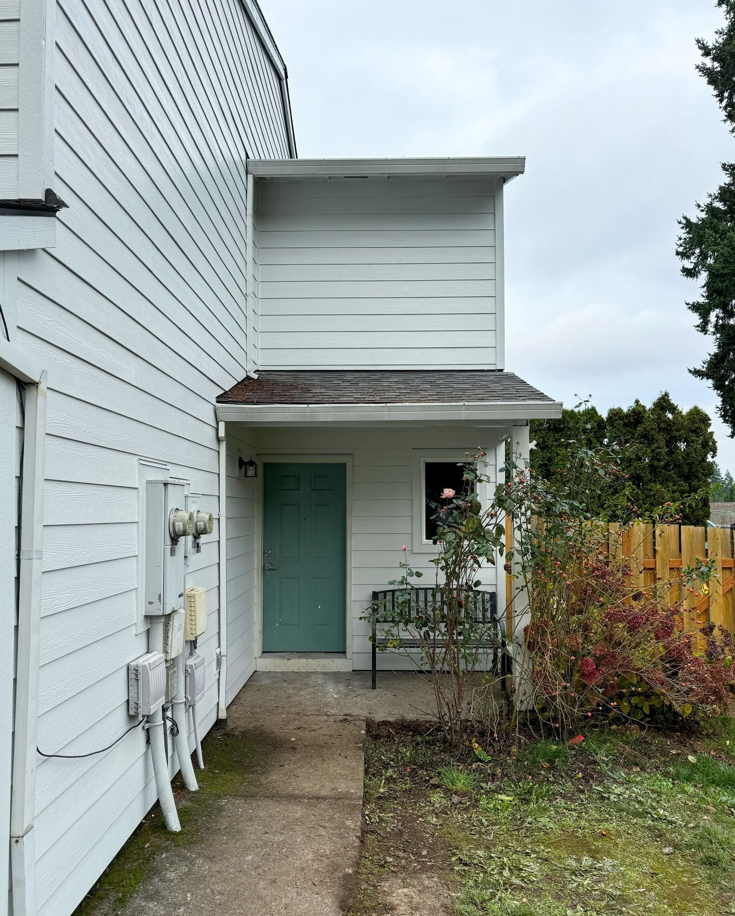 A white building exterior with a teal door, a small porch, a wooden bench, and a concrete walkway.