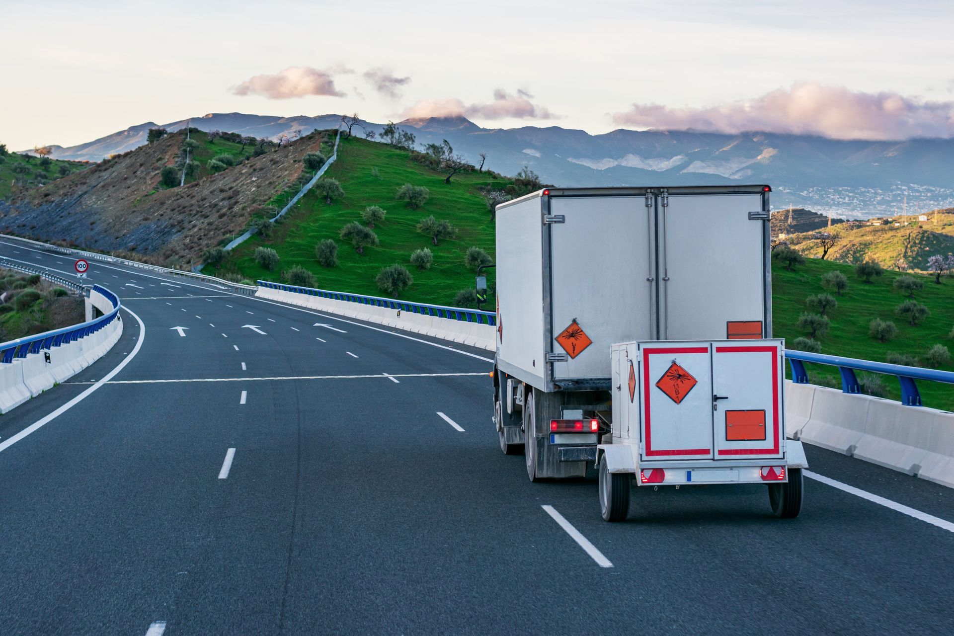 Un camión con remolque que transporta materiales peligrosos circula por una autopista con colinas verdes al fondo.