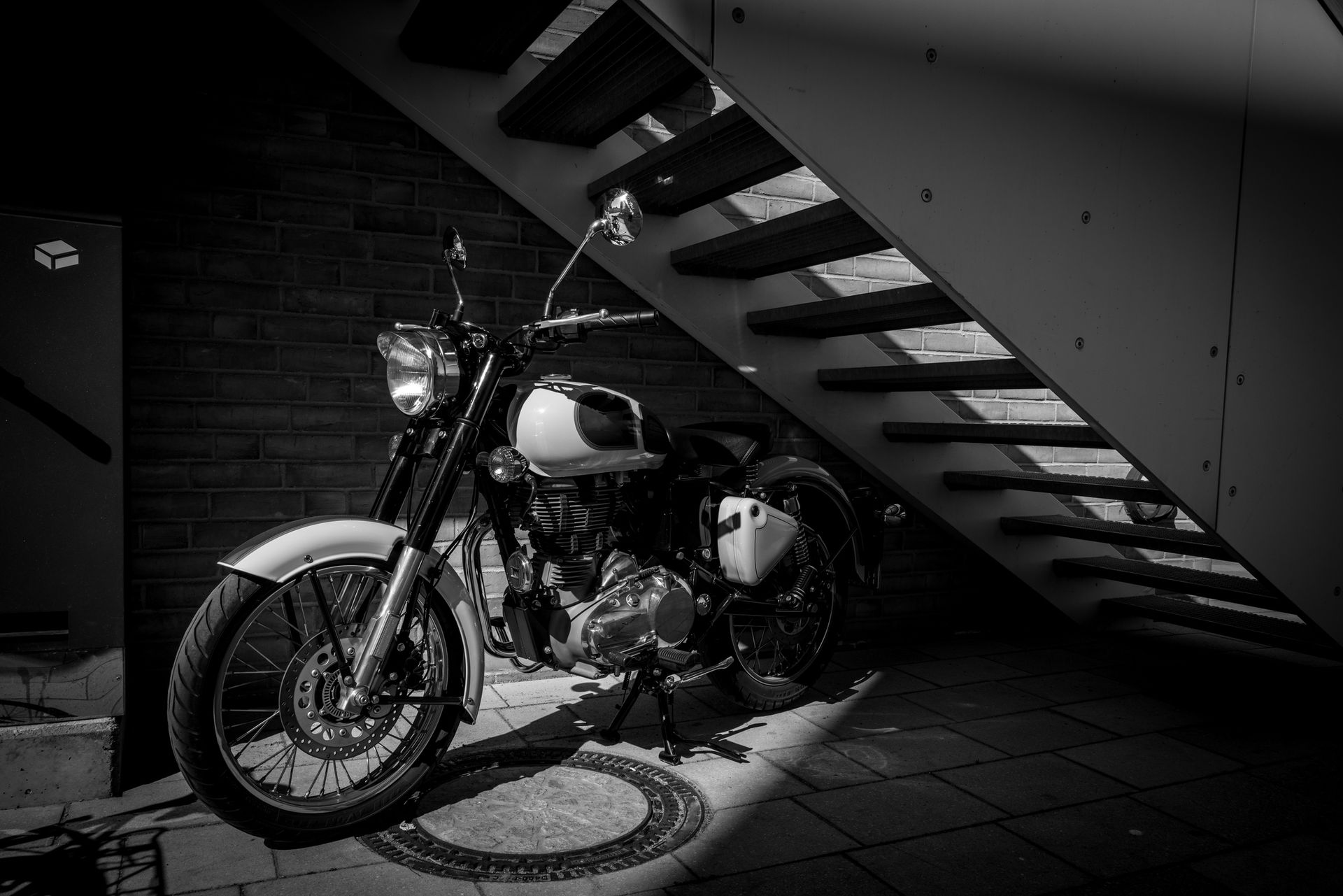 Fotografía en blanco y negro de una motocicleta estacionada debajo de una escalera, iluminada por la luz del sol.