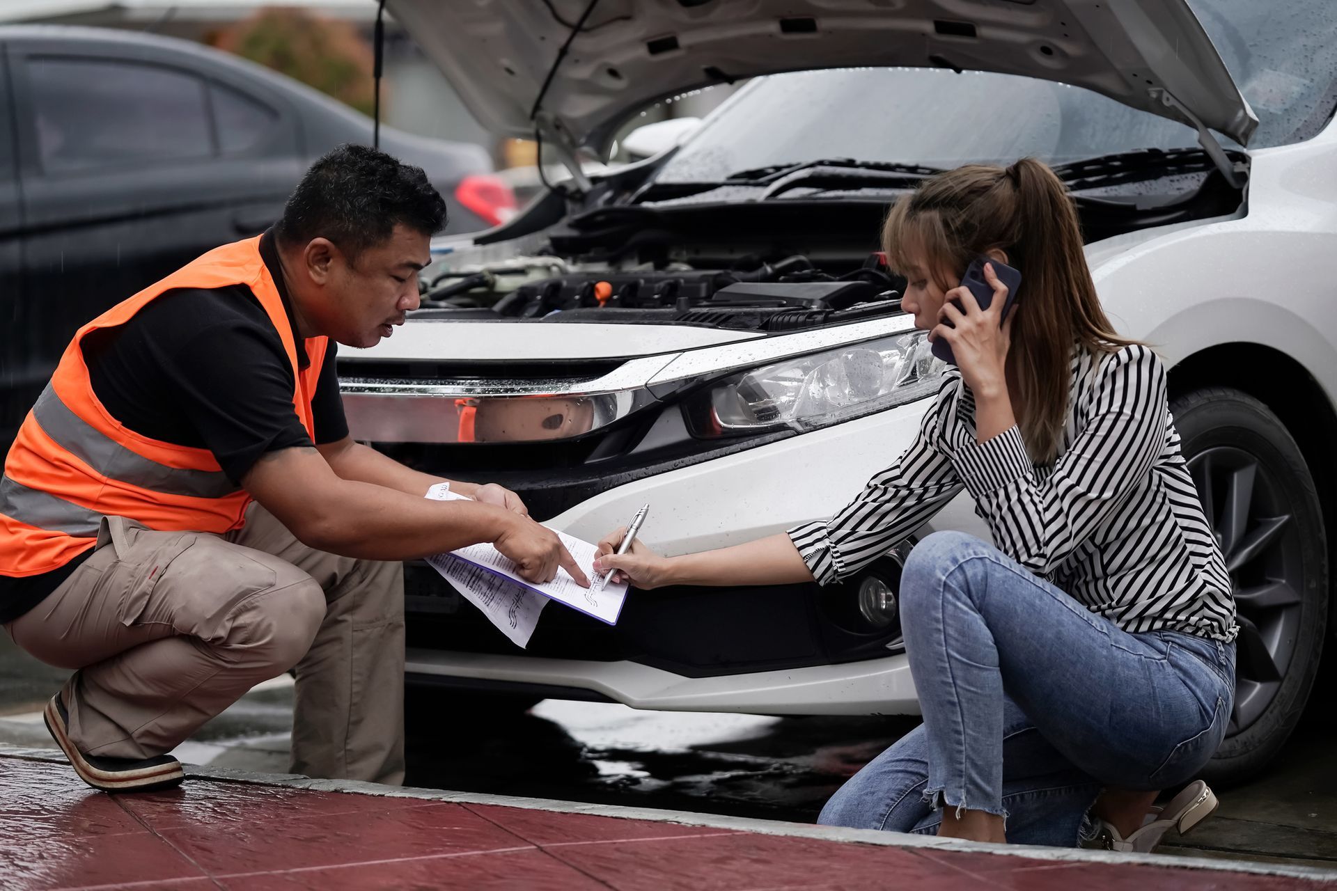 Hombre y mujer en coche con el capó abierto, mujer en el teléfono firmando papeles.