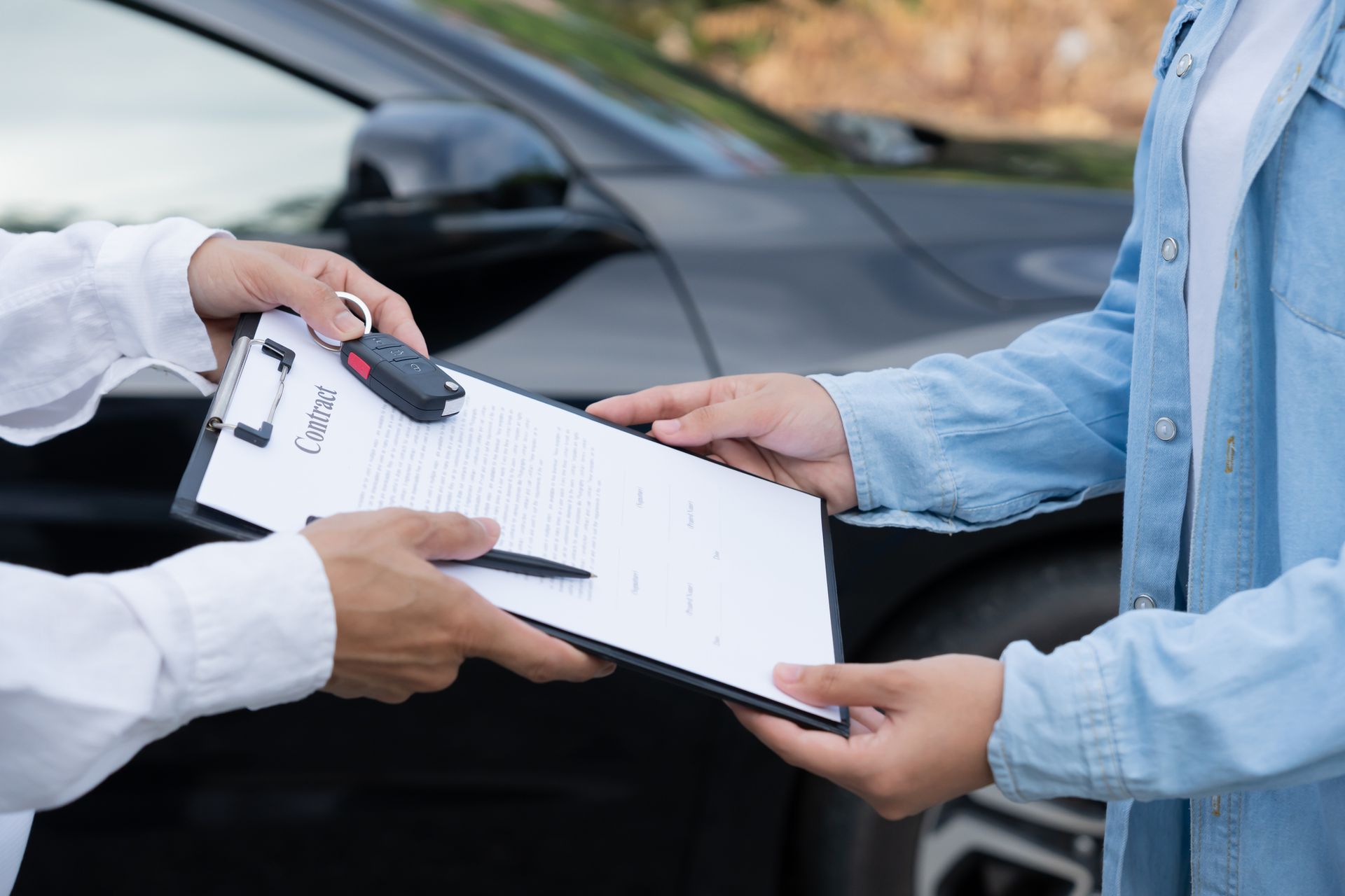 Hands exchanging car keys and contract in front of a black vehicle.