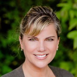 Smiling woman with two-toned dark and blonde hair wearing a grey blazer against a neutral backdrop.