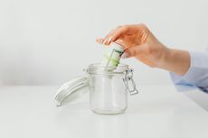 Hand placing rolled cash into an open glass jar on a white table