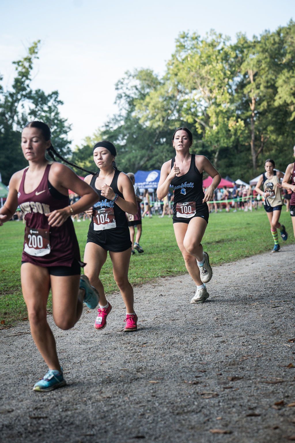 Runners in a cross-country race on a gravel path near trees and a crowd, with bib numbers visible