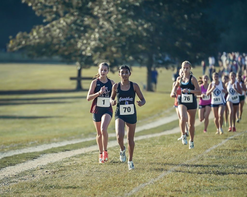 Cross-country runners racing across a grassy field, with two leading the pack and others trailing behind.
