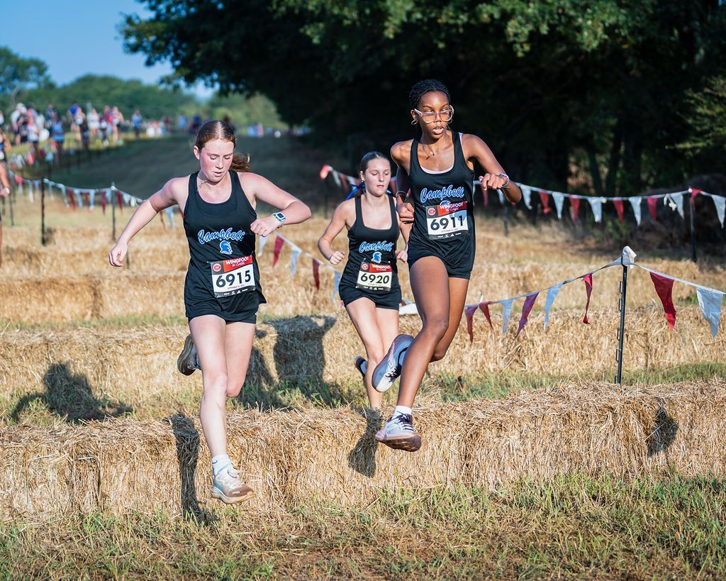 Three runners in black uniforms race through a grassy cross-country course with colorful pennant flags.