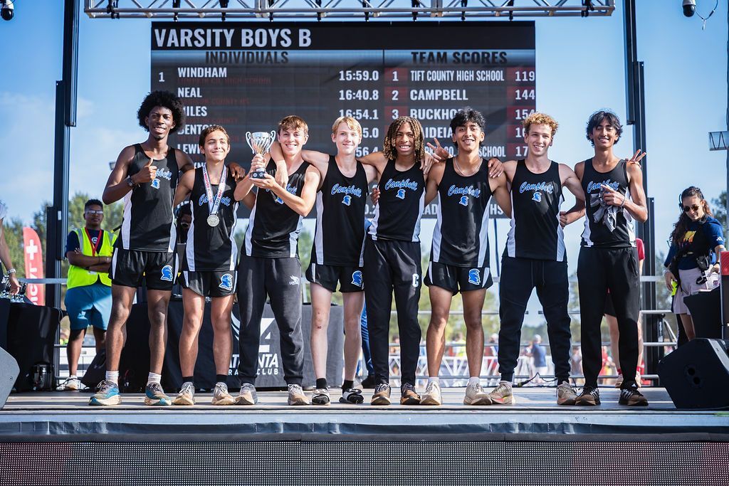 Cross-country team on a podium at a varsity boys B race, wearing black uniforms and raising trophies.
