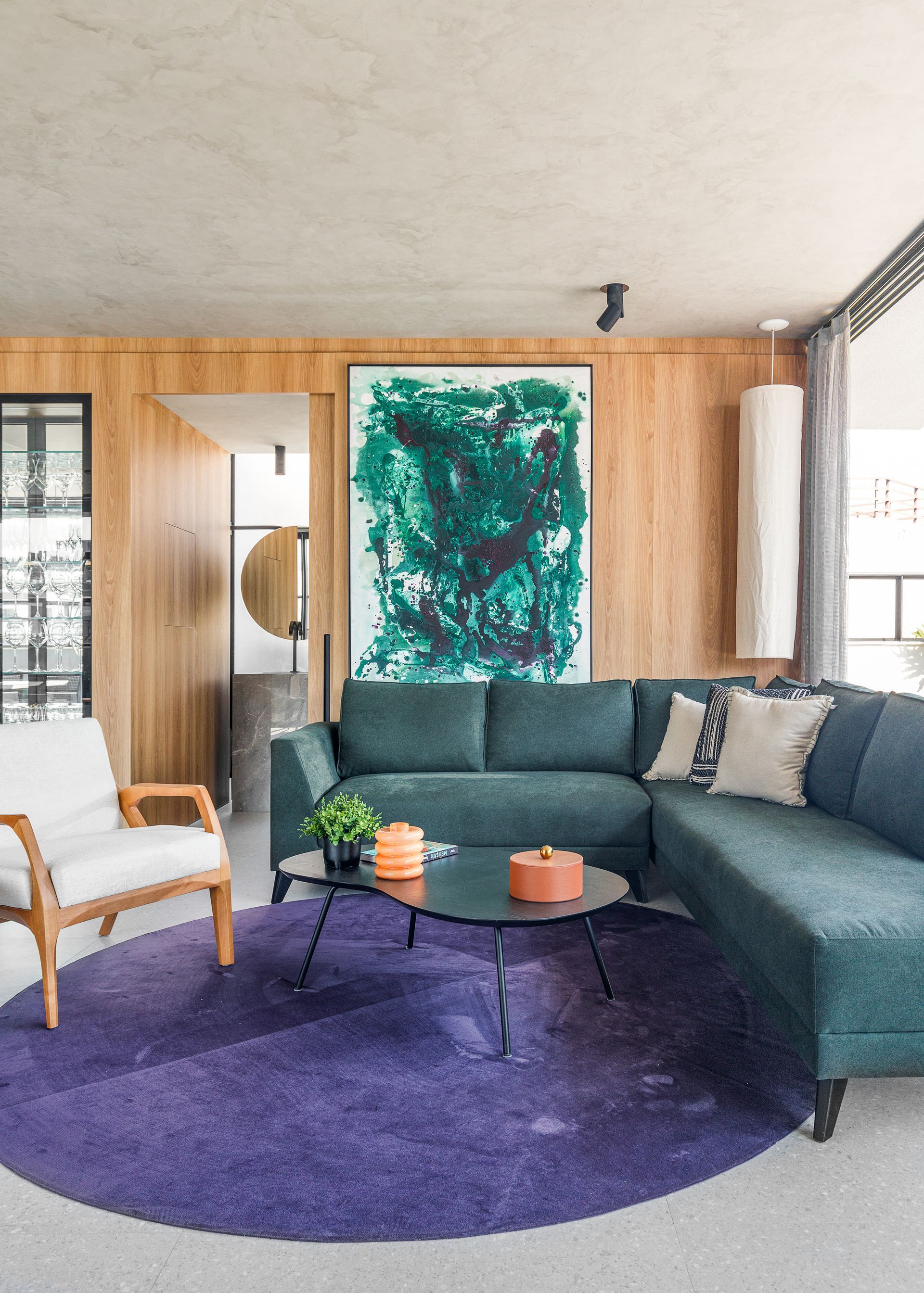 Sala de estar com sofá secional azul-petróleo, tapete roxo e pintura verde abstrata em parede de painel de madeira.