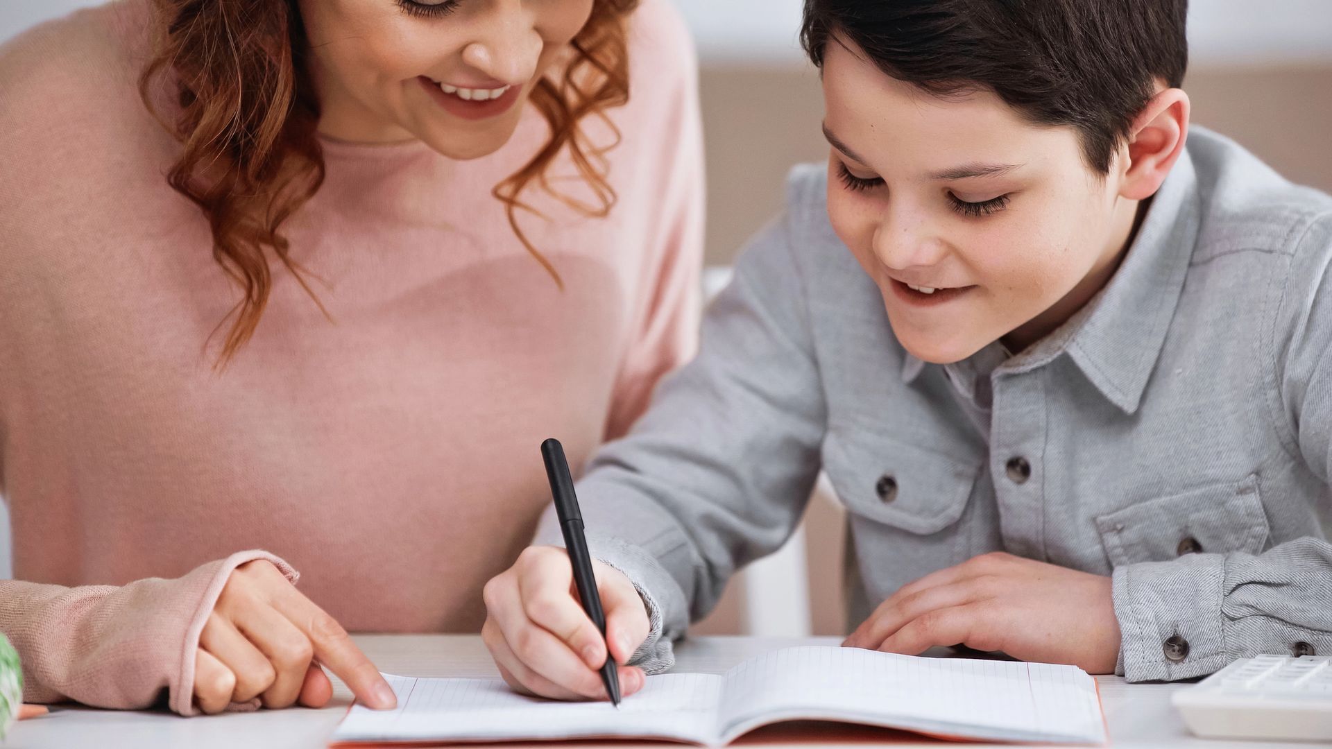 Una donna aiuta un bambino a scrivere su un quaderno; entrambi sorridono.