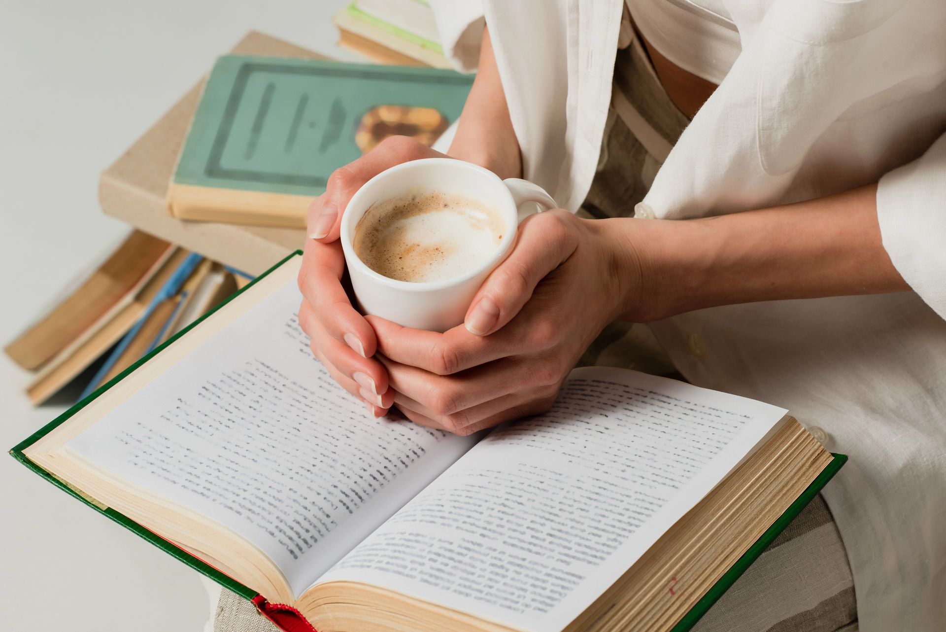 Persona che tiene in mano una tazza di caffè, legge un libro e ha una pila di libri nelle vicinanze.