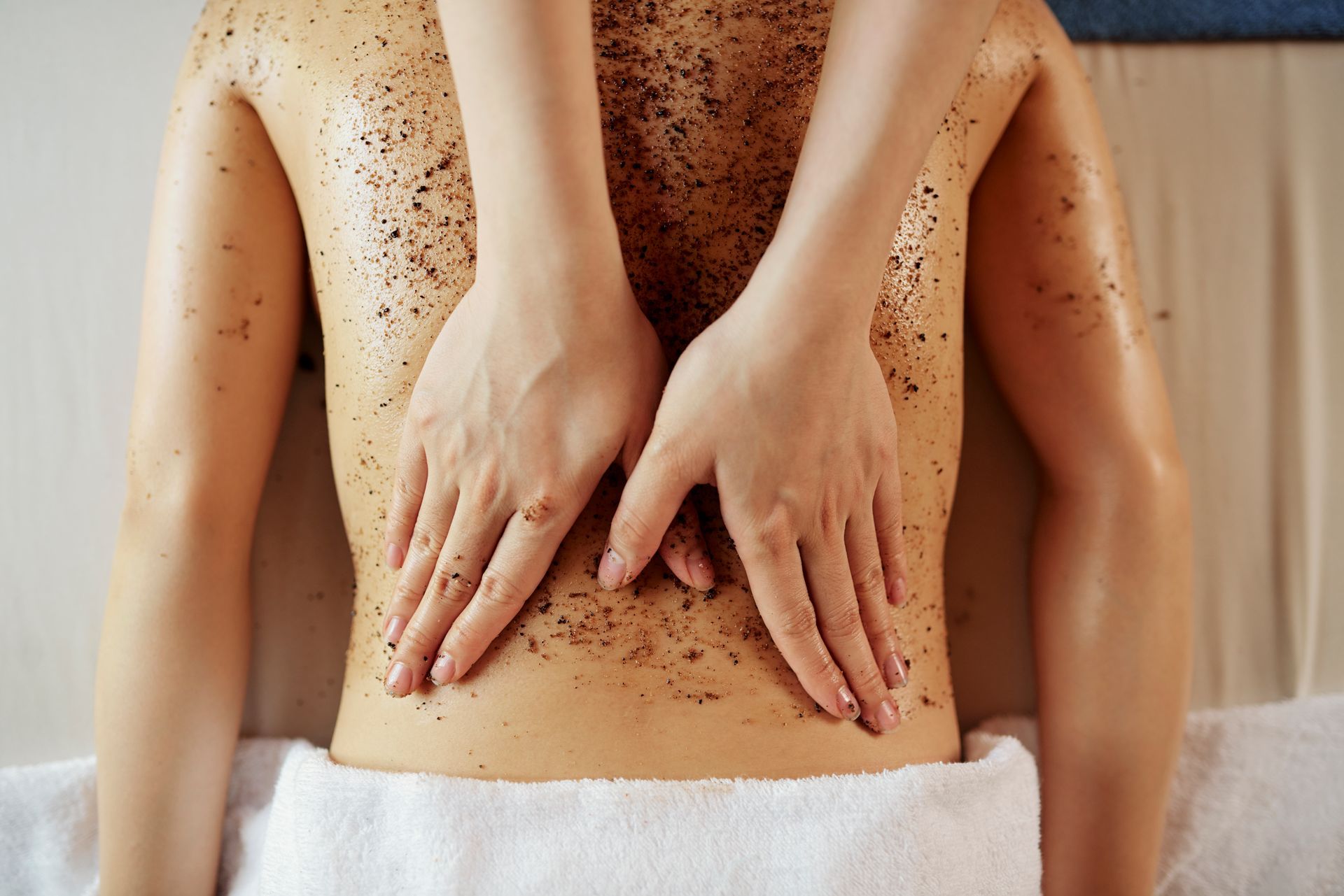Mani che massaggiano la schiena di una persona ricoperta da uno scrub marrone, in una spa.