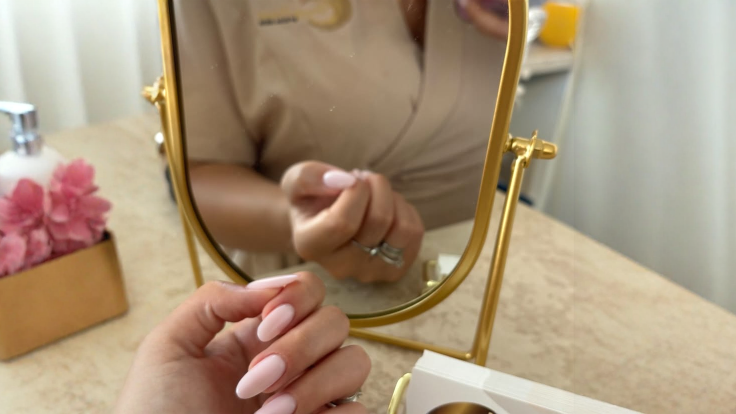 Una donna ammira le sue unghie curate color rosa chiaro in uno specchio con cornice dorata in un salone di bellezza.