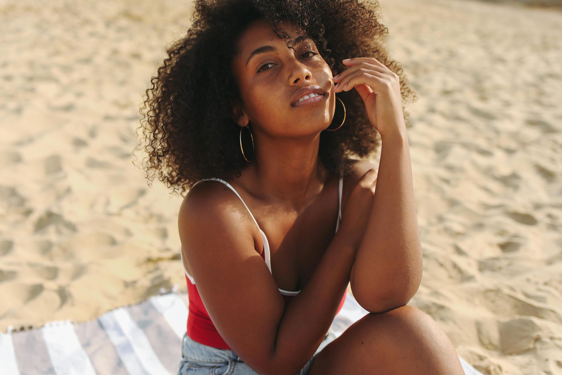 Woman with curly hair sits on beach, wearing red top and denim shorts, resting chin on hand.