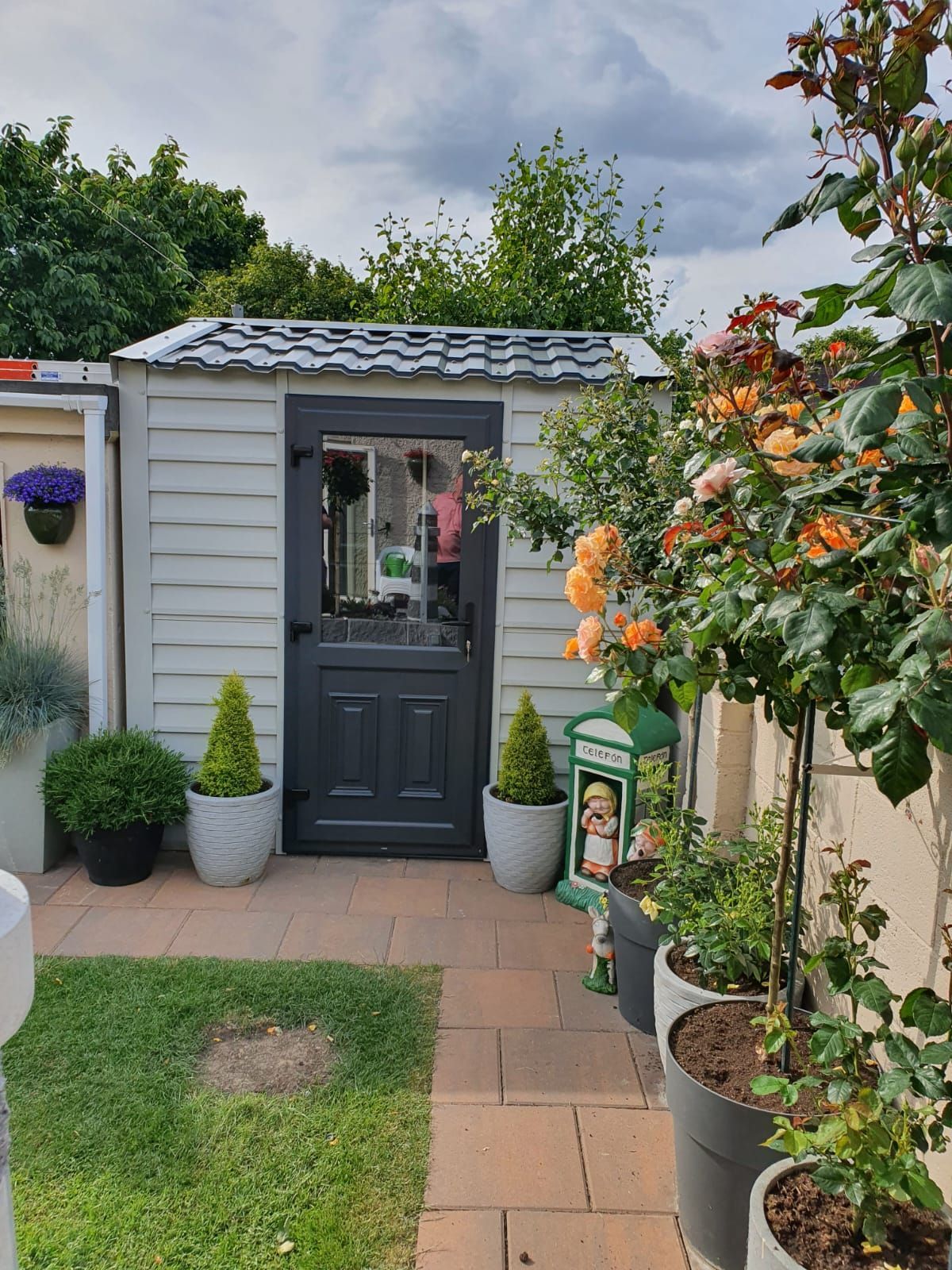 A white shed with a black door is surrounded by potted plants.