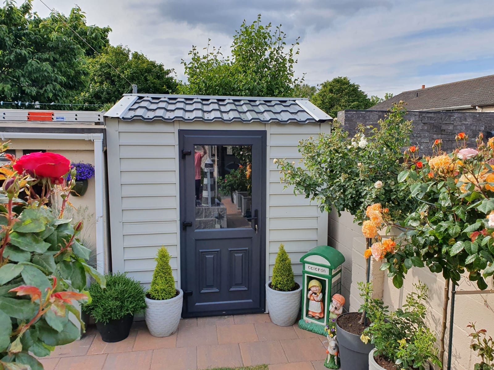 A white shed with a black door is surrounded by potted plants and trees.