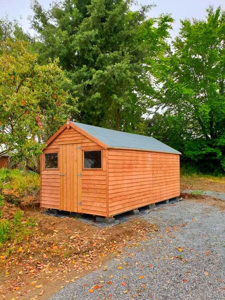 A wooden shed with a blue roof is sitting in the middle of a gravel driveway.