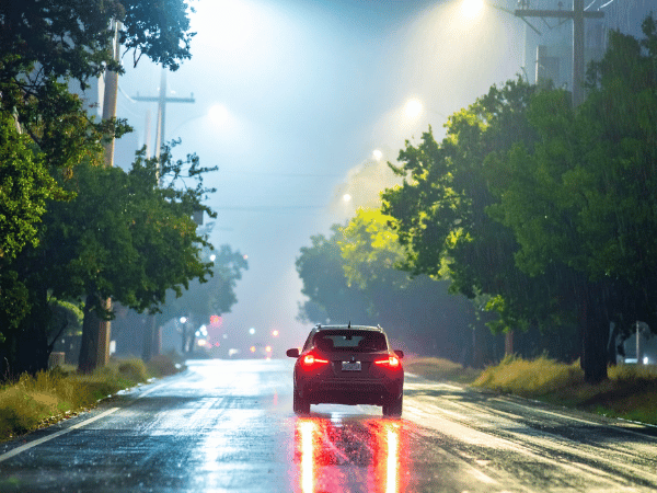 Car driving on a wet road at night with rain falling and taillights reflecting on the pavement under streetlights.