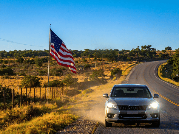 Car parked along a rural road with headlights on and an American flag waving beside the highway.