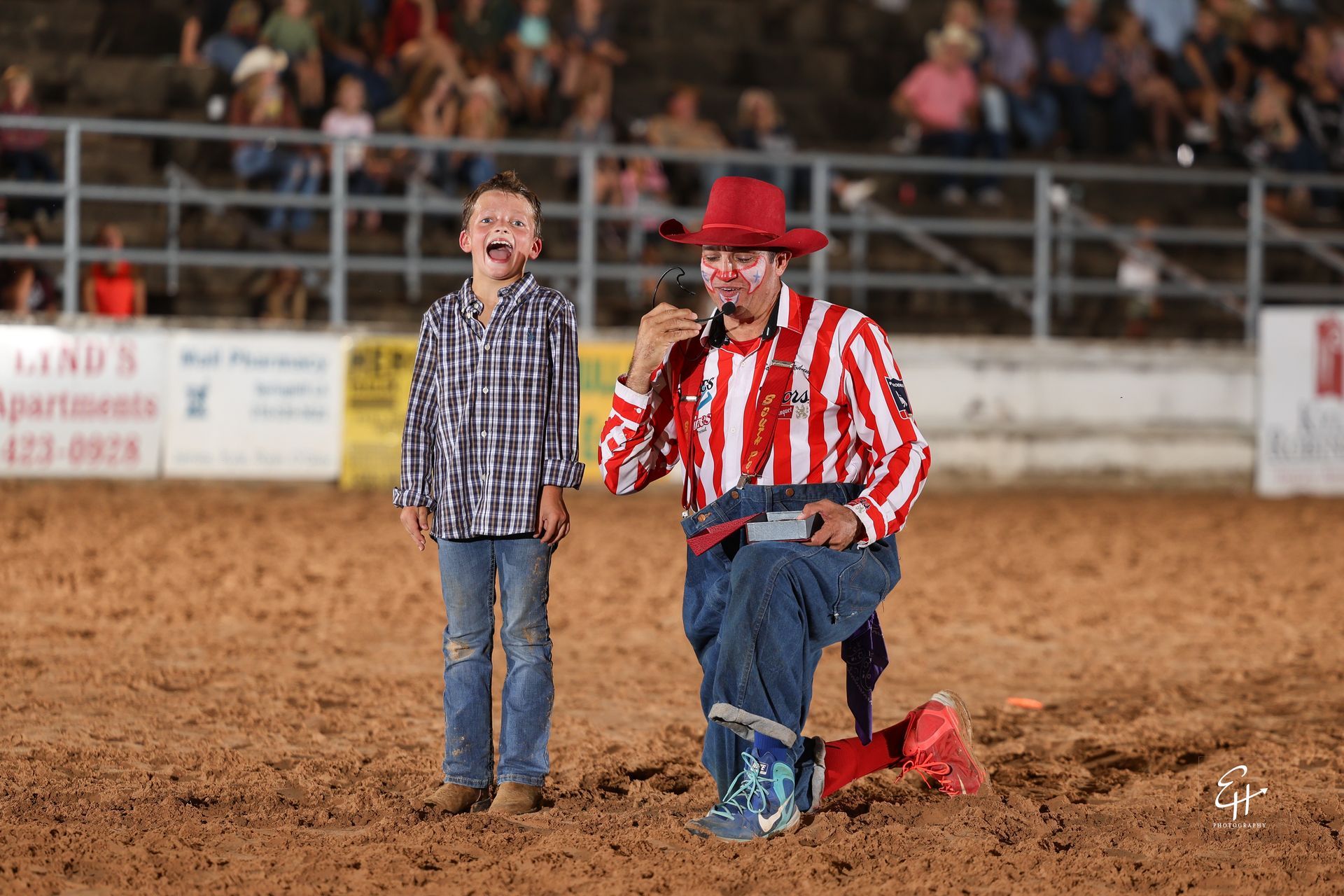 Two people with microphones in a rodeo arena; one kneels in a red striped shirt and hat.
