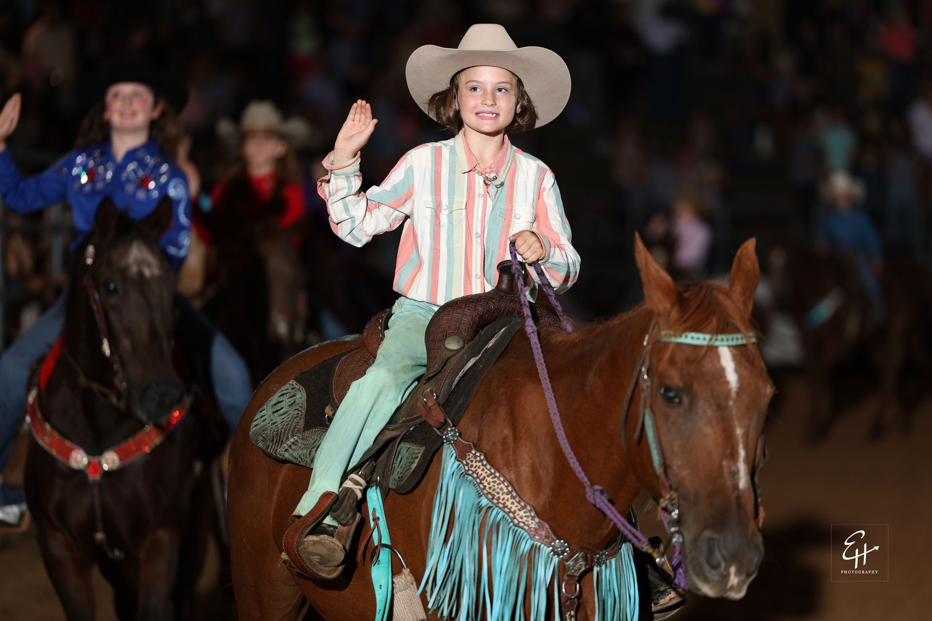 Cowboy in a white hat rides a brown horse in a dim arena, raising a hand in a striped shirt.