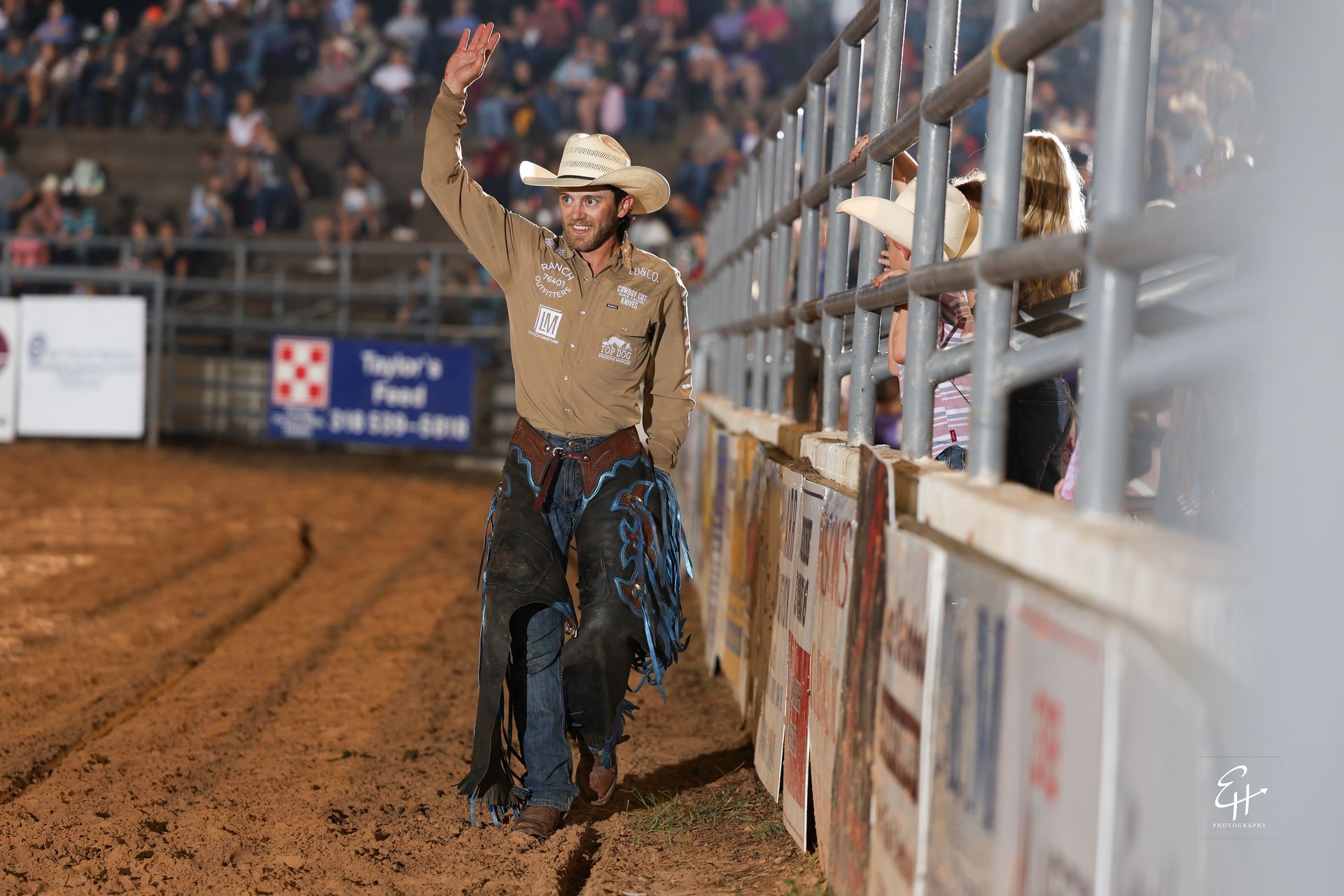 Cowboy in a hat leans on a rodeo arena fence, raising one arm after a ride.