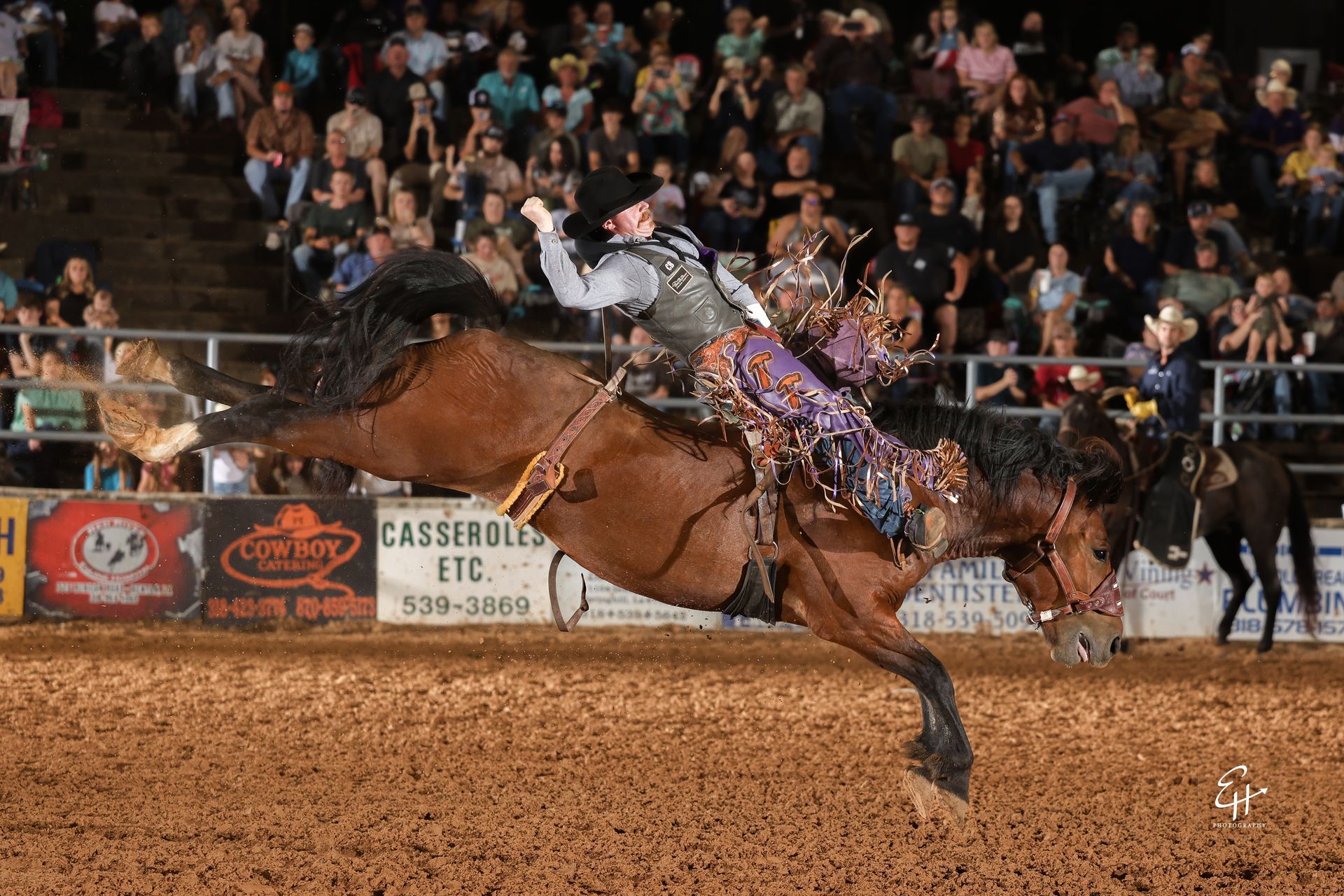 Rodeo rider on a bucking horse in an indoor arena, with a crowd watching in the background.
